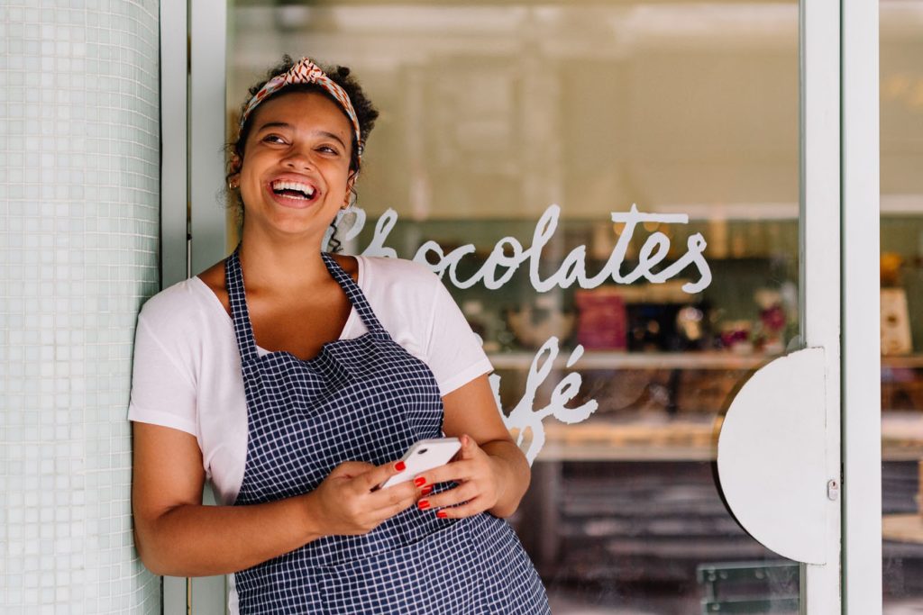 Successful cafe owner, a young woman, stands in her restaurant using a smartphone. Happy female entrepreneur using technology to manage her small business efficiently.