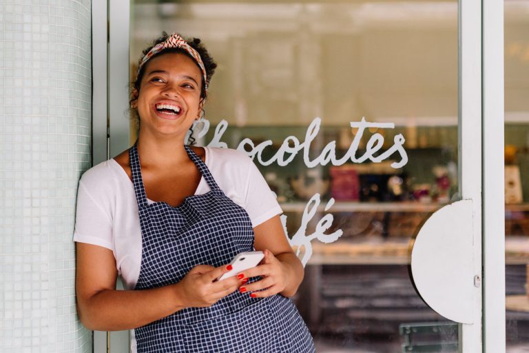 Successful cafe owner, a young woman, stands in her restaurant using a smartphone. Happy female entrepreneur using technology to manage her small business efficiently.