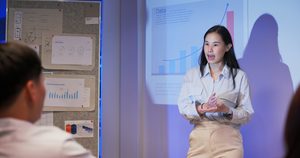 Professional Asian businesswomen lead data-driven presentations with global market insights in modern dark office. Team listens to financial reports, projections, and strategy under projector light.